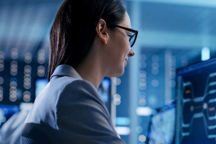 A woman in business attire with glasses is focused on monitoring data in a network server room.